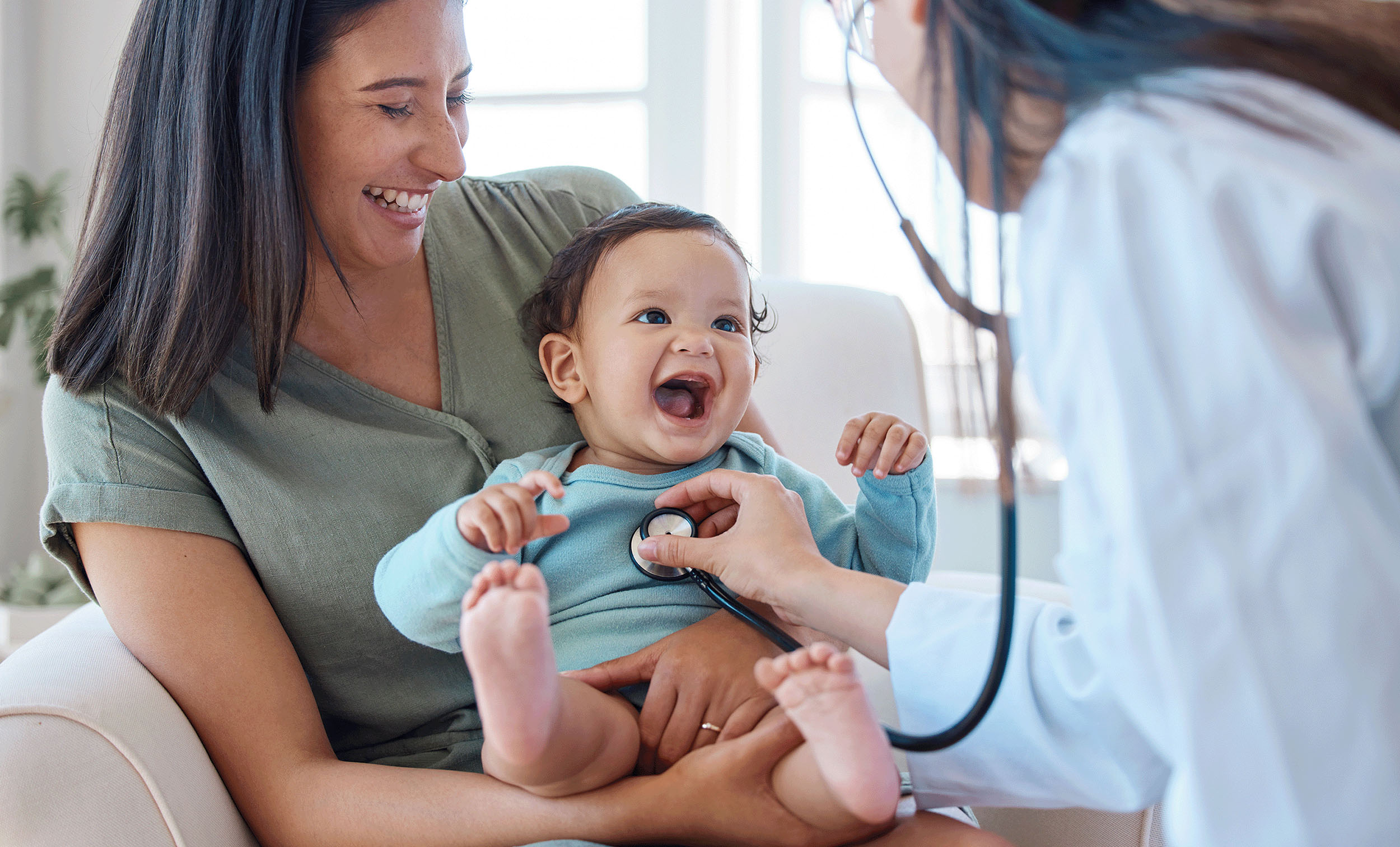 A joyful moment captured as a woman smiles while holding a baby, who is laughing gleefully. A doctor gently places a stethoscope on the baby's chest, symbolizing a heartwarming and reassuring medical examination.