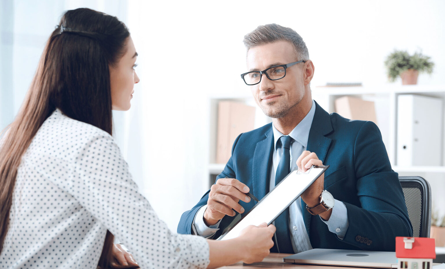 A man in a suit and glasses handing a clipboard to a woman, illustrating a professional interaction and the importance of documentation and communication in a business environment.