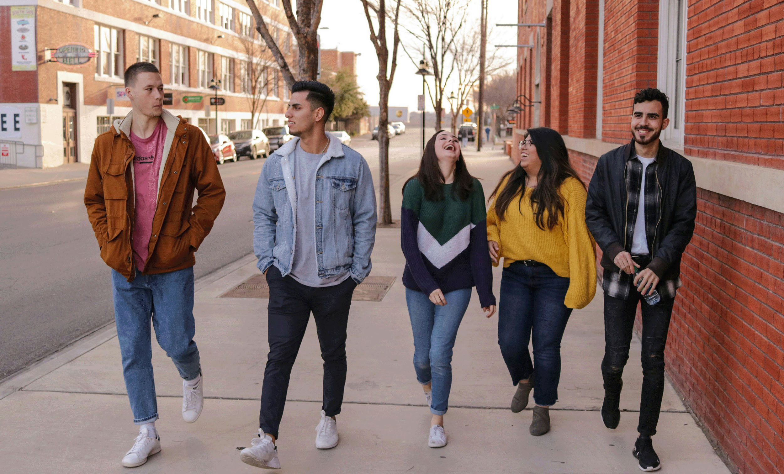 Five young adults walk down a sidewalk next to a brick building. Four of them are conversing and smiling. The weather appears mild.