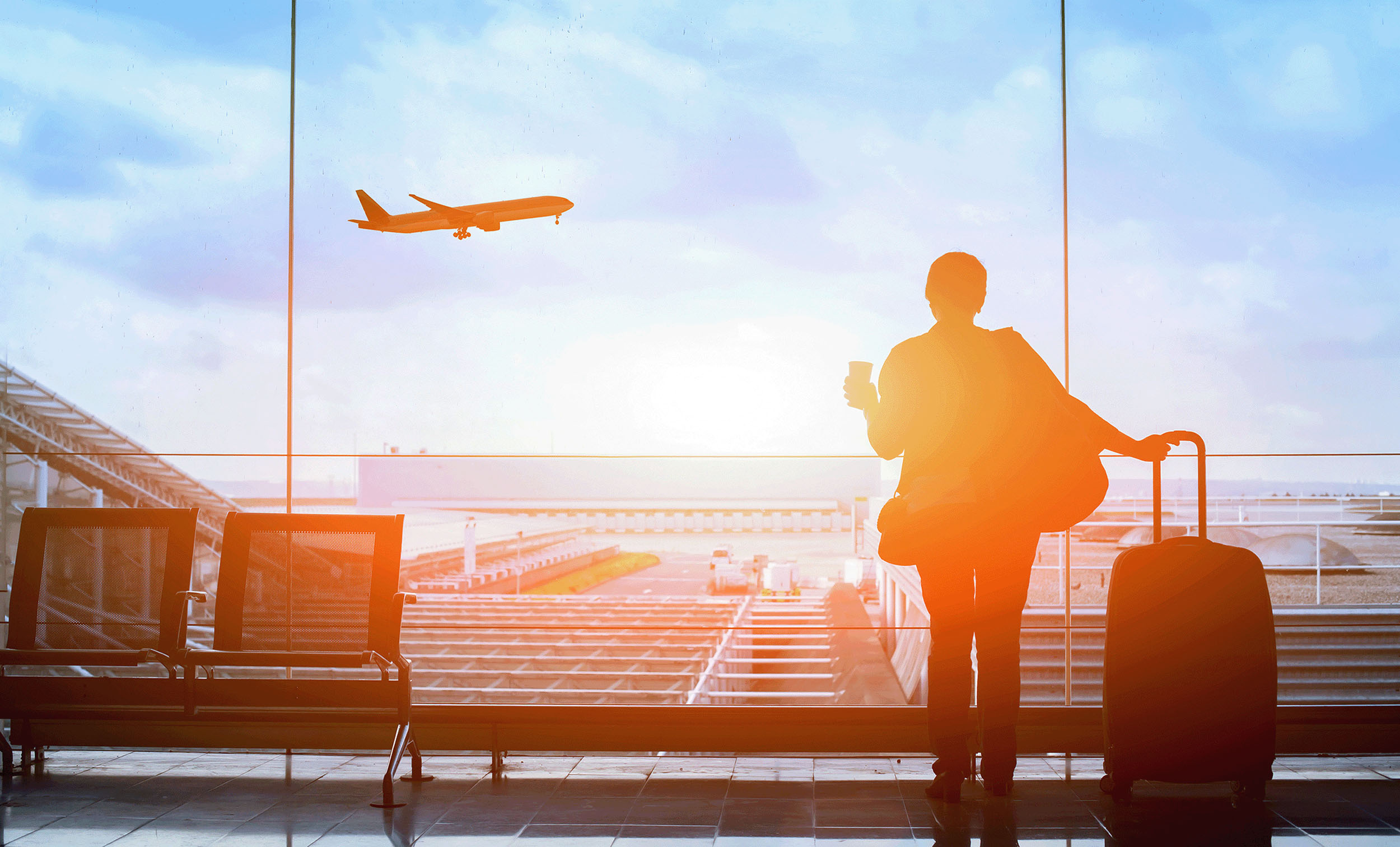 A person at an airport gate looks out at a plane taking off while holding a cup and a rolling suitcase.