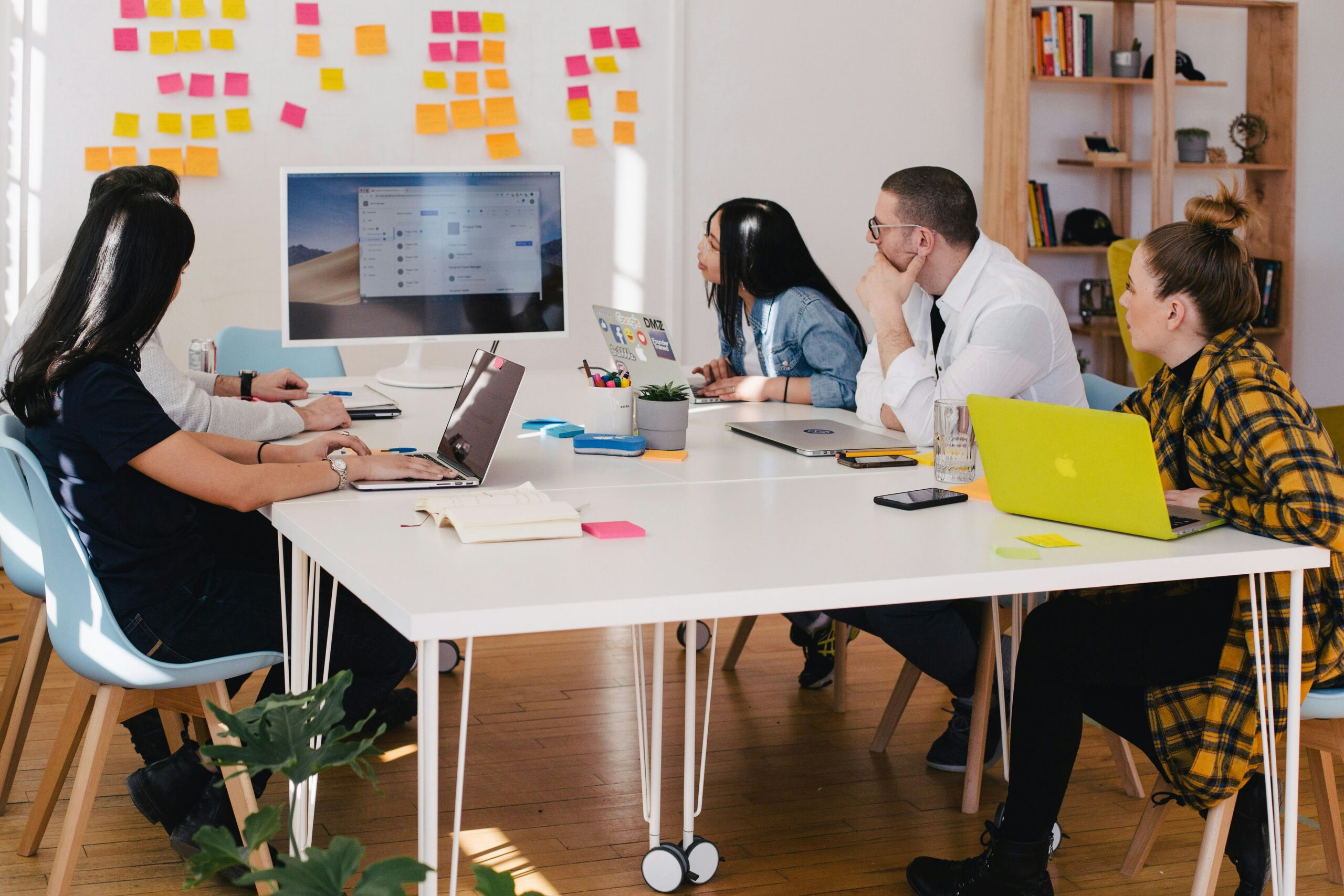 A diverse group of five people engaged in a meeting around a large white table, with laptops and notepads in front of them. The room is decorated with colorful sticky notes on the wall and shelves with books and plants